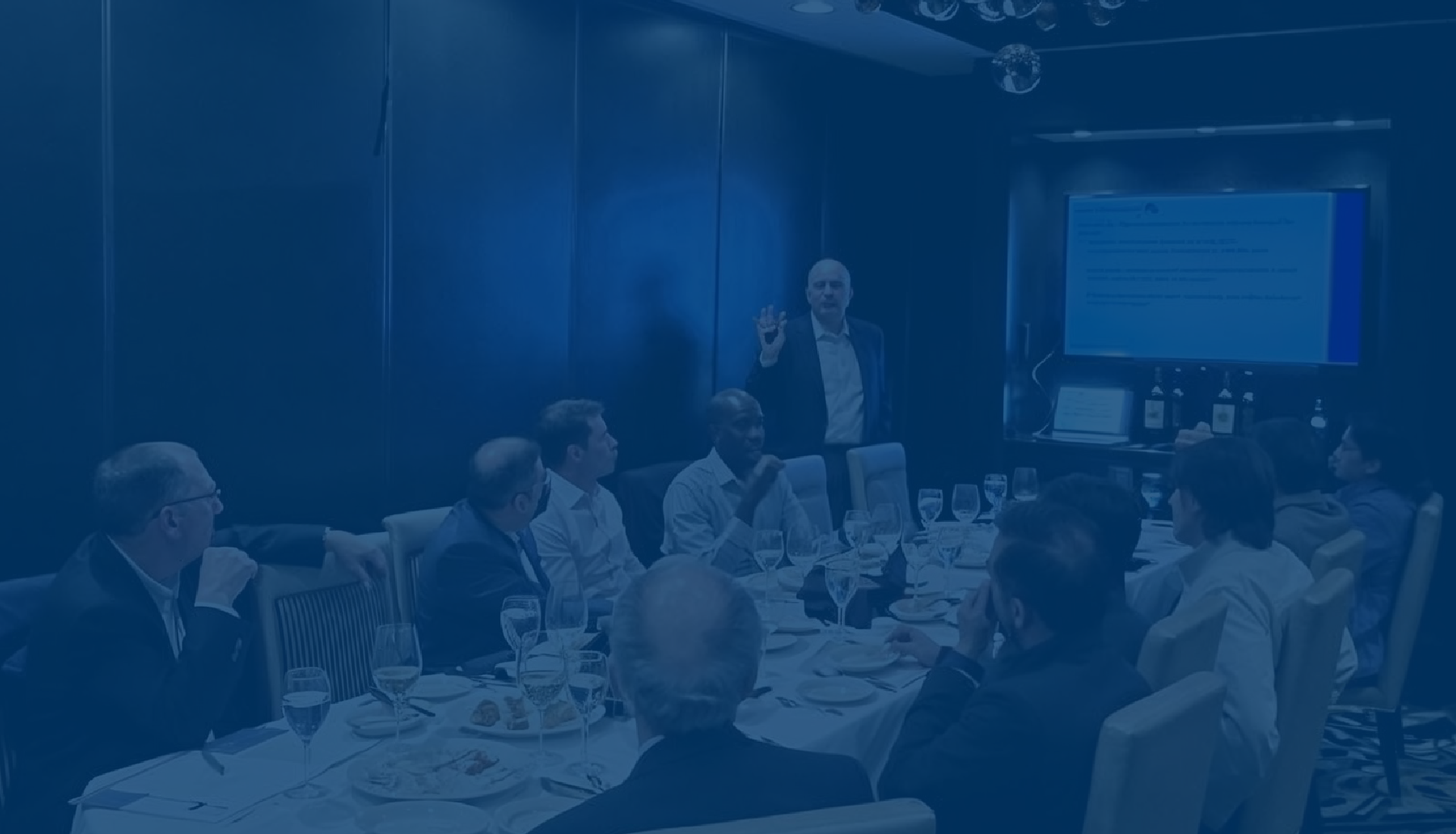 A group of people seated around a long dining table in a private event room, looking towards a large projection screen displaying a presentation. The tables are set with food and drinks. There is a presenter at the front of the room leading the presentation.